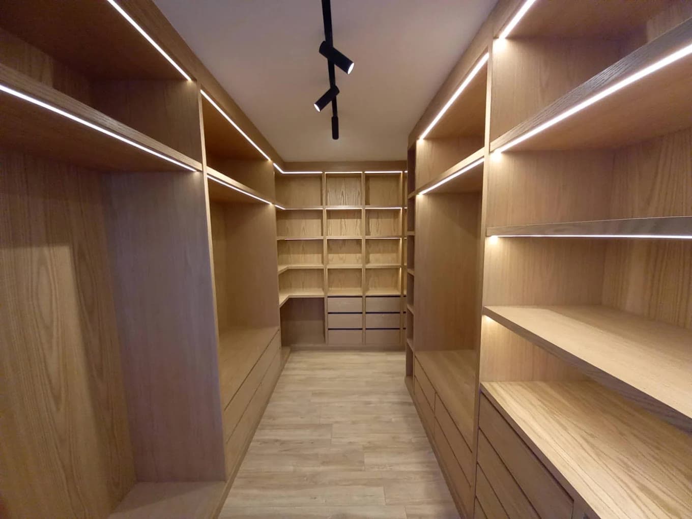 Dressing en bois clair en forme de couloir, avec étagères, tiroirs et éclairage linéaire sous les tablettes