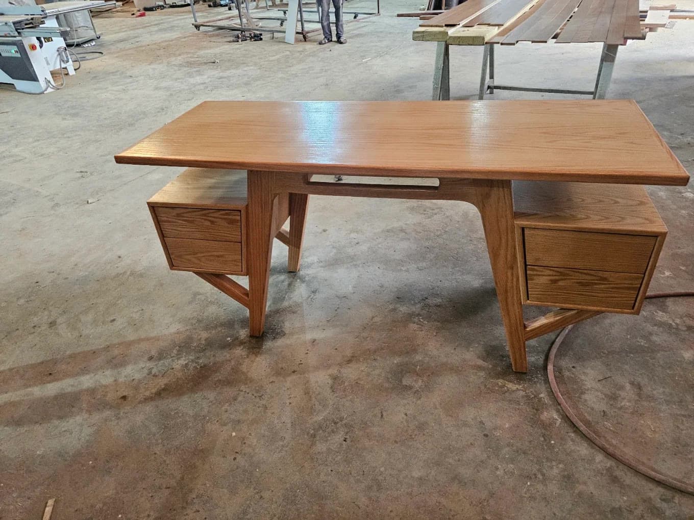 Bureau en bois à deux caissons de tiroirs sur pieds inclinés, vu de face dans l’atelier avec des panneaux sur un établi